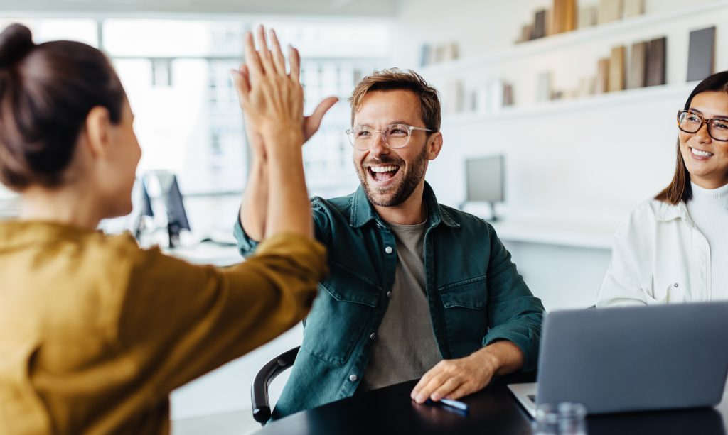 Fröhliche Büroszene: Zwei Personen geben sich ein High-Five, während eine weitere Person daneben sitzt und lächelt.