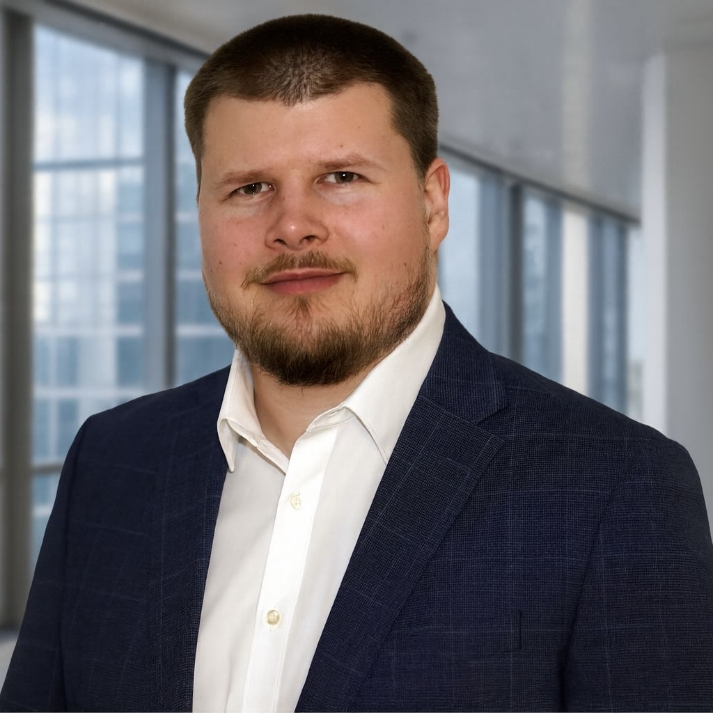 Ein Mann in einem blauen Businessanzug und weißem Hemd steht in einem modernen Büro mit großen Fenstern, durch die eine Stadtlandschaft mit Hochhäusern zu sehen ist.