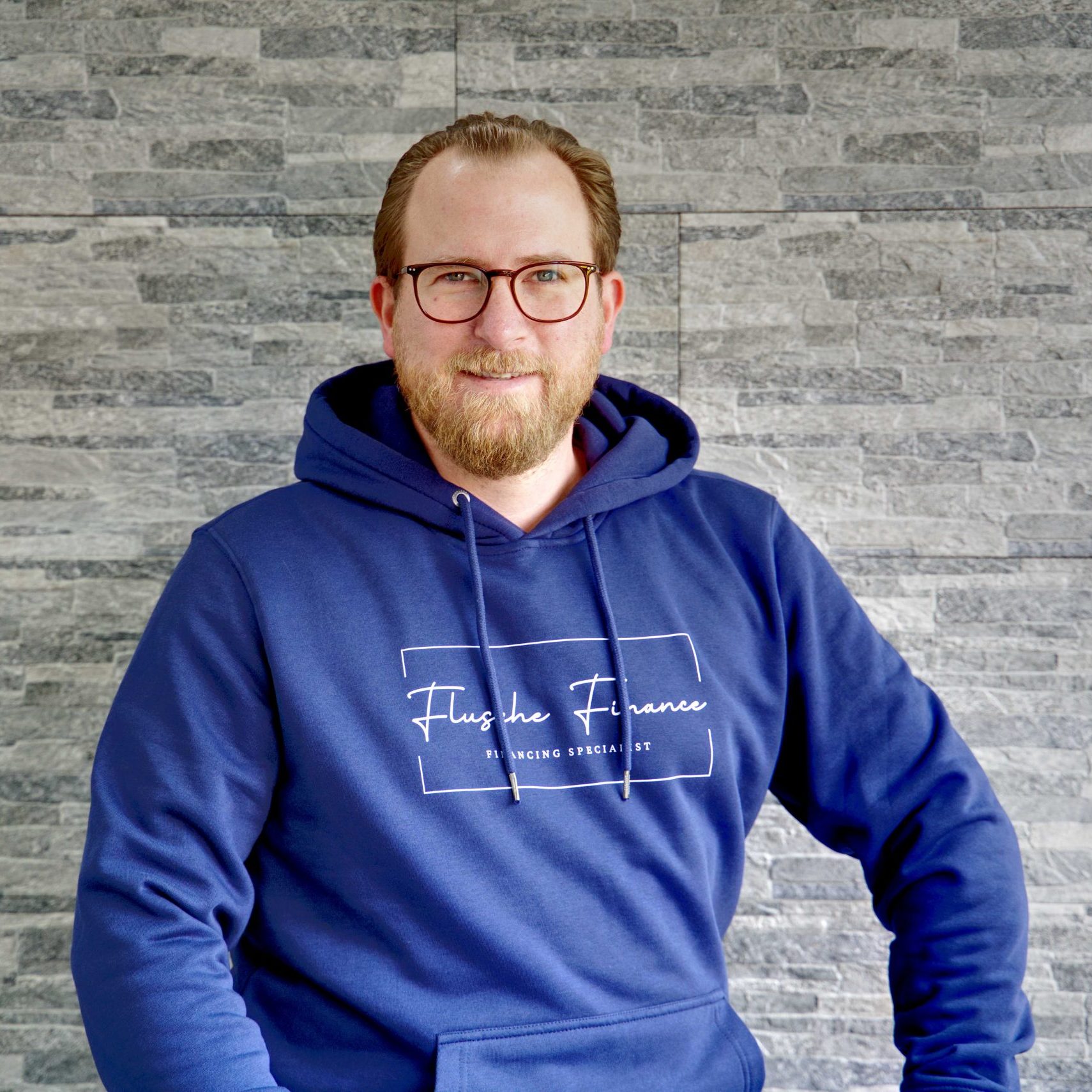 Bild mit Inhaber. Lächelnde Person mit blauem Sweatshirt und Brille vor grauem Hintergrund.